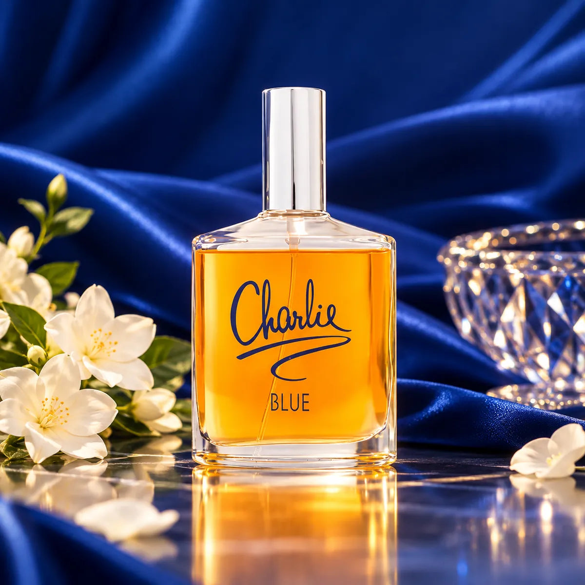 Frasco de Charlie Blue de Revlon con líquido ámbar, flores blancas, tela azul y reflejos sobre superficie brillante 